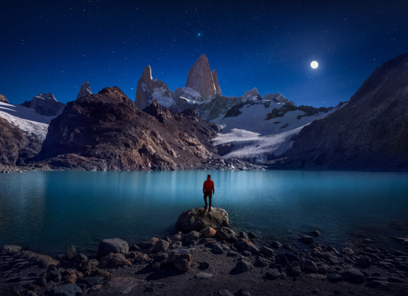 A person in a red jacket stands on a rock by a tranquil, reflective lake surrounded by jagged, snow-capped mountains under a starry night sky with a bright moon.