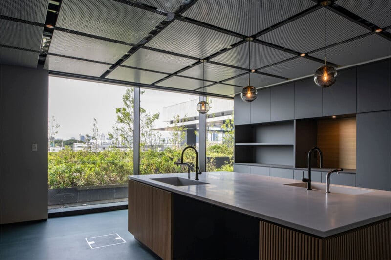 A modern kitchen with a large island featuring two sinks and minimalist cabinetry. The room has ceiling-mounted light fixtures and a large floor-to-ceiling window with a view of outdoor greenery. The sleek design elements emphasize clean lines and ample natural light.