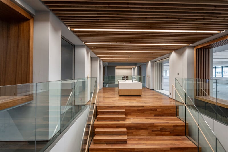 A modern, open office space with wooden floors and stairs. The ceiling has exposed wooden beams, and large windows provide natural light. Glass railings line the stairs and adjacent walkways, leading up to a central desk area with a minimalist design.