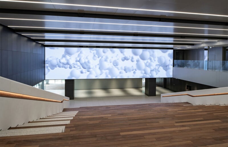 A modern, wide staircase with wooden steps and white railings leads down to an open area. A large digital screen displaying a pattern of white bubbles on a light background spans the wall in front of the stairs. The space is illuminated with ceiling lights.