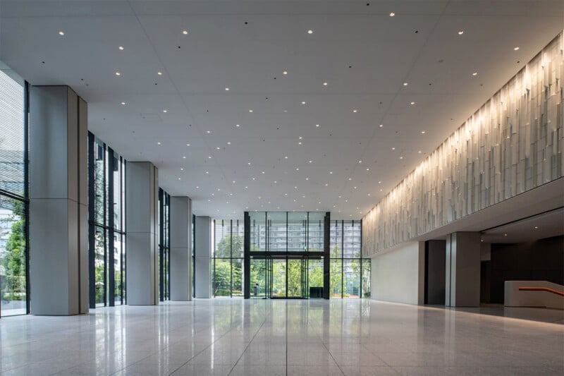 A spacious, modern lobby with a high ceiling and large glass windows. The space is well-lit by numerous recessed lights on the ceiling. The floor is covered in polished tiles, and the windows provide a clear view of greenery outside, creating an open, airy atmosphere.