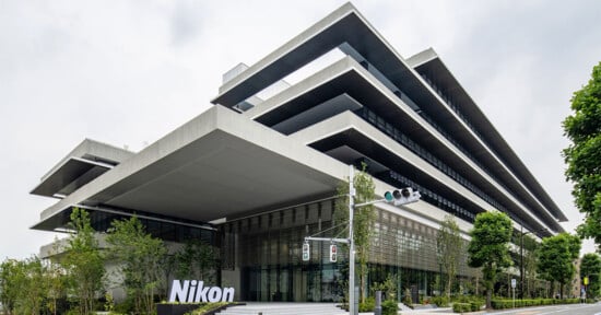 A modern, angular building with multiple overhanging floors, surrounded by greenery and trees. The building has a sign reading "Nikon" at its entrance, and traffic lights are visible in the foreground. The sky is cloudy.