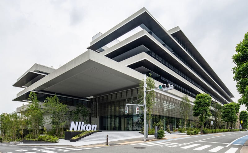 Modern building with multiple staggered, overhanging floors and extensive use of glass and angular design features. The Nikon logo is prominently displayed at ground level. The foreground includes trees, a pedestrian crosswalk, and traffic lights. Sky is overcast.