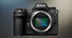 A close-up of a Nikon Z series mirrorless camera with no lens attached. The camera body is black and features various labeled buttons, a textured grip on the left side, and a prominent electronic viewfinder at the top. The sensor is visible in the lens mount.