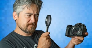 A man with gray hair and a beard is holding a camera in his left hand and inspecting it closely with a magnifying glass in his right hand. He has a serious and focused expression. The background is blue with a pixelated pattern.