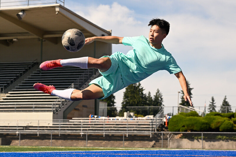 A young soccer player in a turquoise uniform is captured mid-air executing a dynamic kick with a soccer ball. The stadium stands and field are visible in the background, along with clear weather and trees. The player displays intense focus and athleticism.