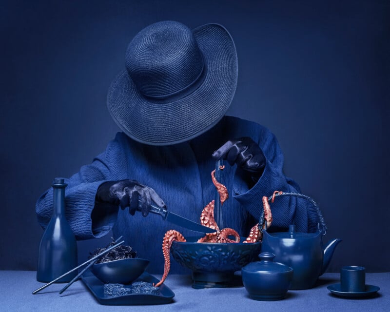 A mysterious figure in a blue outfit, including a wide-brimmed hat and gloves, is seated at a blue-draped table. They are using a fork and tongs to handle a piece of octopus, surrounded by matching blue crockery and utensils. The entire scene has a monochromatic blue theme.