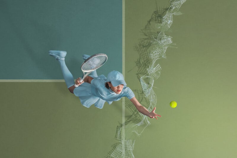 A female tennis player, dressed in light blue, is captured mid-air as she extends her arm to hit a yellow tennis ball with her racket. The action takes place on a green and blue tennis court with a broken net visible below her.