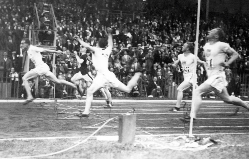 A black and white photo of a track and field race finish line. Several male runners are captured mid-stride as they cross the tape. The crowd in the background appears engaged, watching the thrilling conclusion of the race.