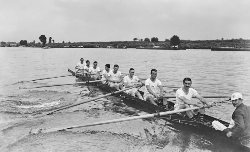 A black and white photo showing an eight-man rowing team on the water, all in light-colored shirts, synchronized as they row. A coxswain, seated at the stern in a dark jacket and cap, guides the team. The background features a calm waterway and a distant shoreline.