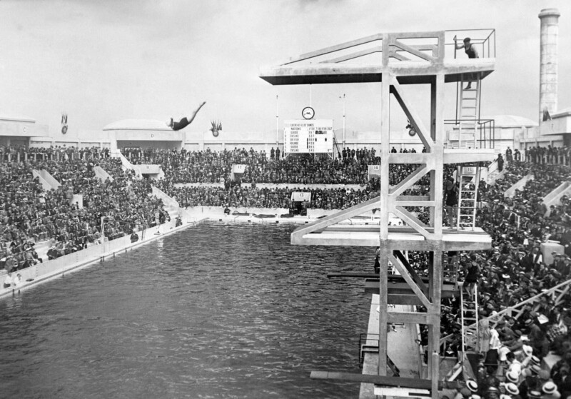 A black-and-white image of a crowded outdoor diving arena. A diver is captured mid-air above the pool, having just jumped from a high diving platform. Spectators fill the stands surrounding the pool, and a large scoreboard and clock are visible in the background.