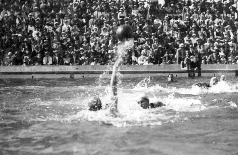 A black-and-white photo showing a water polo match in progress. Players are in the water, reaching for the ball. In the background, a large crowd of spectators is seated, closely watching the game. The scene appears lively and competitive.