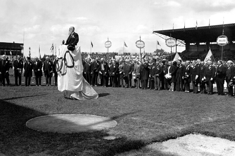 A man stands on a platform draped with an Olympic flag, addressing a large crowd gathered on a grassy field with a stadium backdrop. People surround the platform, many wearing suits and hats. Banners displaying country names are visible in the background.