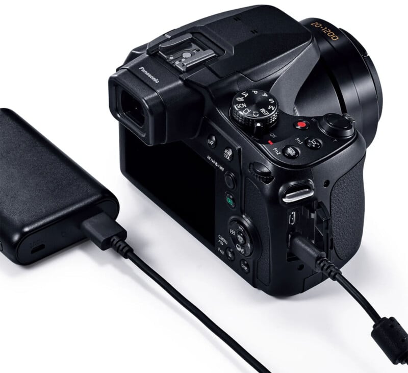 Close-up of a black digital camera connected to an external power source via a USB cable. The camera features various buttons, dials, and a viewfinder. The black power adapter is plugged into an electrical outlet.