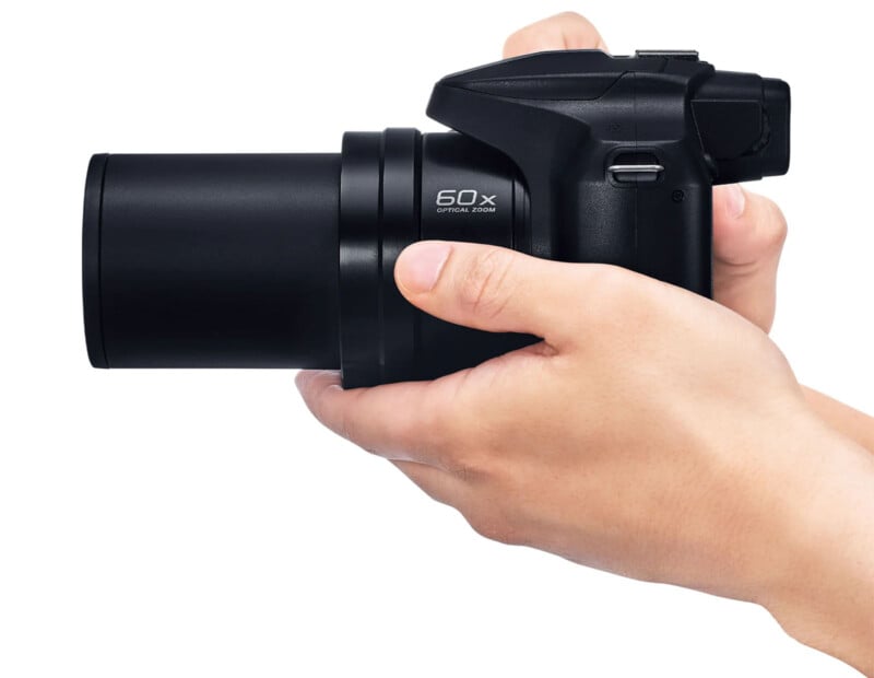 A person holding a black digital camera with a zoom lens pointing to the left. The camera has "60x Optical Zoom" written on the side. The person's right hand is gripping the camera, with the index finger on the shutter button.