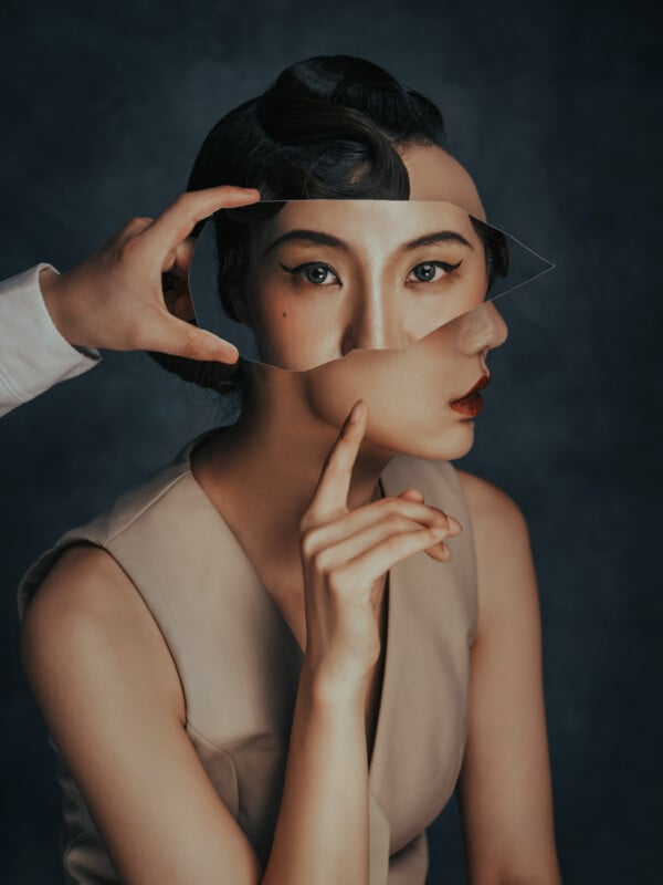 A person with a unique hairstyle holds a jagged, reflective fragment in front of their face, causing parts of their face to appear distorted. The background is a muted, dark blue, and the individual is wearing a sleeveless, beige outfit. The overall tone is surreal.