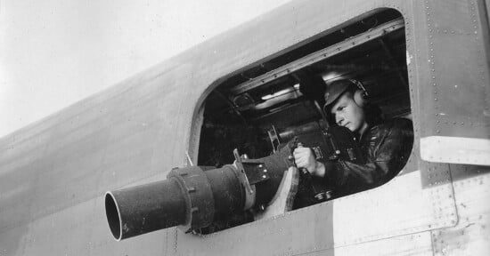 A person in a military uniform and helmet is manning a machine gun mounted at a window of an aircraft. The individual is focusing intently on the weapon, which protrudes outside the aircraft's body, suggesting a wartime scenario.