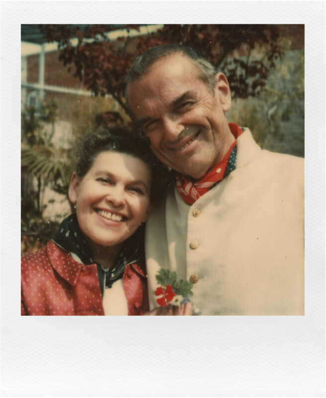 A smiling couple stands closely together outdoors. The woman has curly hair and wears a red jacket with a white scarf. The man has short hair and wears a beige jacket with a red bandana around his neck. He has his arm around her shoulders.