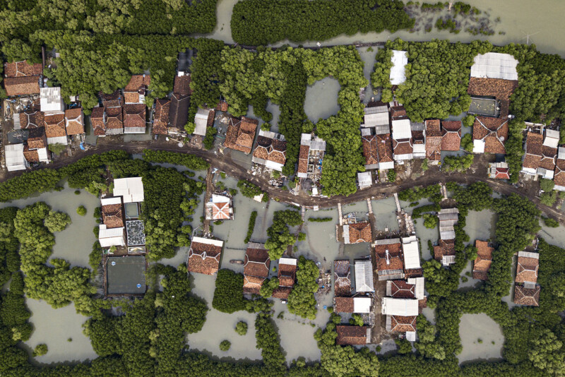 Aerial view of a flood-affected village surrounded by green vegetation. Traditional houses with reddish-brown roofs are partially submerged in water. The flooded area creates a pattern of isolated clusters of homes and green patches.