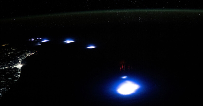 A view of Earth at night from space, showing bright blue bioluminescent spots in the ocean. City lights are visible along the coastline to the left. The dark horizon and numerous stars are visible above.