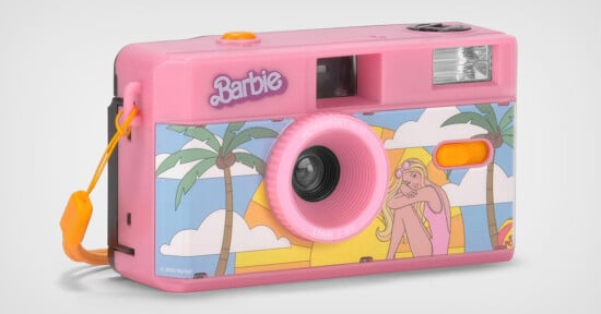 A pink Barbie-themed film camera with a drawing of a girl lounging under palm trees on the front. The camera features a wrist strap and a flash near the top right corner. The Barbie logo is displayed near the top left of the camera.