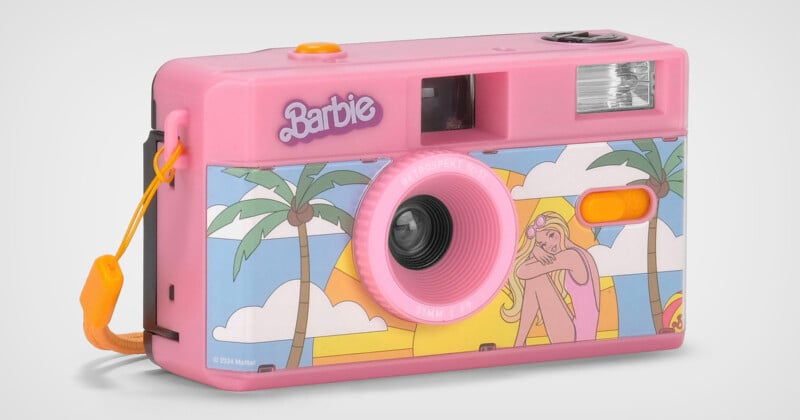 A pink Barbie-themed film camera with a drawing of a girl lounging under palm trees on the front. The camera features a wrist strap and a flash near the top right corner. The Barbie logo is displayed near the top left of the camera.