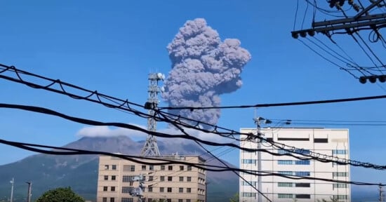 A volcano erupts, sending a large cloud of ash into the clear blue sky. The eruption is visible behind buildings and numerous power lines in the foreground. The scene is framed by urban structures, highlighting the contrast between nature and civilization.