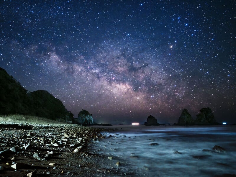 A breathtaking night sky filled with countless stars and the Milky Way galaxy, over a rocky coastline with gentle waves lapping at the shore. Dark silhouettes of cliffs and rock formations can be seen against the horizon, creating a serene and magical scene.