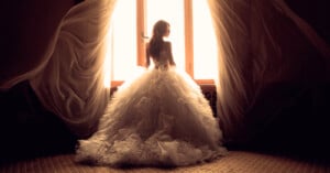 A bride in a voluminous, elegant white gown stands facing a bright window with sheer curtains billowing on either side. The soft lighting creates a warm and ethereal atmosphere, highlighting the layers of her dress which flow to the floor.