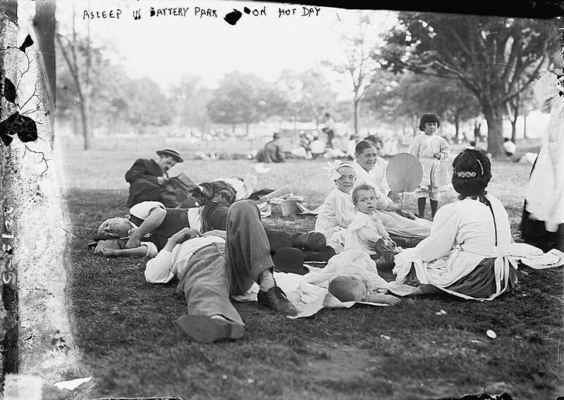 A group of people, including men, women, and children, resting and reclining on the grass in Battery Park on a hot day. Some are lying down while others are sitting upright, and a few are holding fans. Trees are visible in the background.