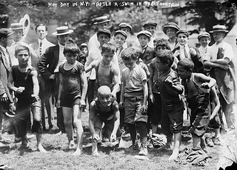 A group of boys, some in swimsuits and others in wet clothes, stand together after swimming in a fountain on a hot day in New York. They are surrounded by a crowd of men and boys wearing hats and suits, some smiling and others looking on. The image is black and white.