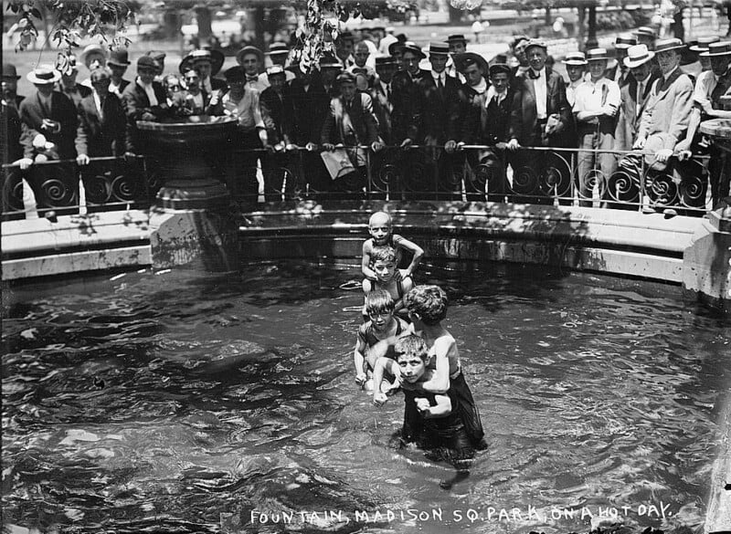 Black and white photo of children splashing in a fountain at Madison Square Park on a hot day, with a crowd of men in suits and hats watching from behind a fence. The scene is lively and captures the relief and joy of cooling off in the fountain.