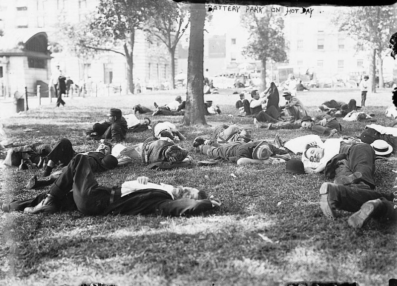 A black and white photo of numerous people lying on the grass in Battery Park, New York, on a hot day. Some are sleeping, while others appear to be resting or relaxing in the shade of various trees. Buildings and a few passersby can be seen in the background.