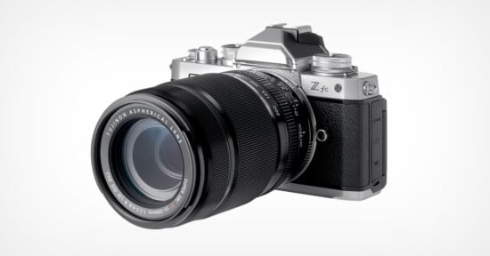 A silver and black Fujifilm digital camera with a large interchangeable zoom lens is placed against a plain white background. The camera features a retro aesthetic with various dials and buttons on the top, including a textured grip on the right side of its body.