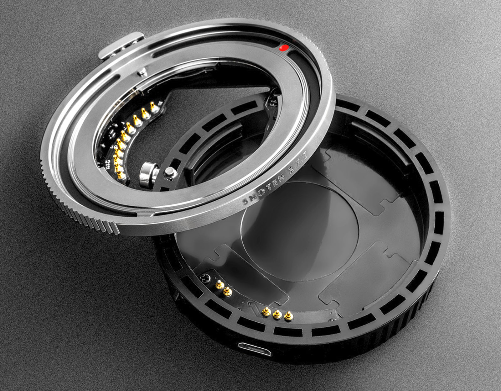 A close-up image of a metal and plastic camera lens adapter, shown opened to display its internal electronic contacts and mechanical components. The adapter has a circular shape with intricate detailing and connectors for interfacing with camera lenses.