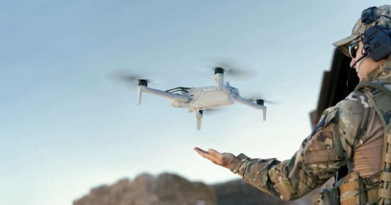 A soldier in camouflage uniform and wearing tactical gear operates a small white drone which hovers above their open hand. The background shows a clear sky and rocky terrain.