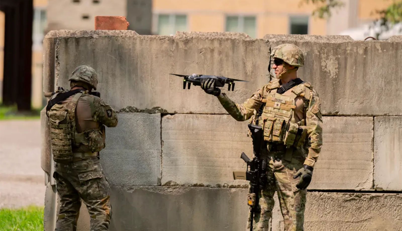 Two soldiers dressed in camouflage uniforms and tactical gear are operating a drone. One soldier is extending his arm to launch or land the drone, while the other is standing with a weapon, facing away, and appears to be inspecting the area. They are near a concrete wall.