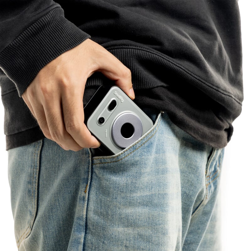 A person wearing a black sweatshirt and jeans slides a gray Instax instant camera into their front jeans pocket.