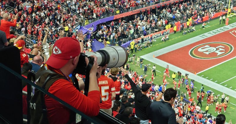 A photographer wearing a Kansas City Chiefs cap and holding a large camera lens takes photos from the stands while the crowd celebrates in the background during a football game. Players and personnel are visible on the field.