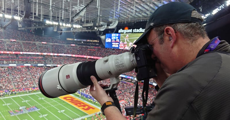 A photographer wearing a cap and lanyard uses a large telephoto lens to capture images at a football game at Allegiant Stadium. The field is visible in the background along with stadium lights and a full audience in the stands.