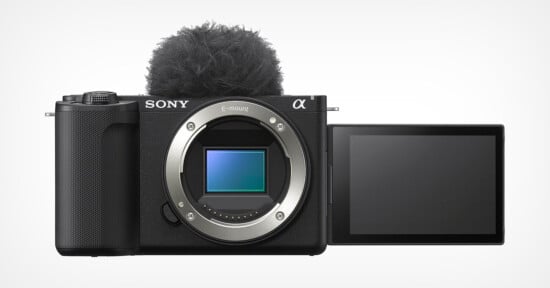 A Sony mirrorless camera with the E-mount lens removed, revealing the sensor. The camera has a flip-out LCD screen on the right, currently positioned outward. A fuzzy windscreen is attached to the top, suggesting it's set up for video recording.