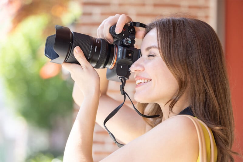 A woman with light brown hair is smiling as she holds a camera up to her face, taking a photo. She is wearing a yellow top and appears to be outdoors, with a blurred background that includes greenery and a brick wall.