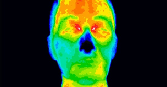 A thermal imagery of a human face displaying heat distribution with varying colors: red and orange for high heat areas, yellow and green for moderate heat, and blue for cooler areas. The background is black, highlighting the contrasted thermal colors.