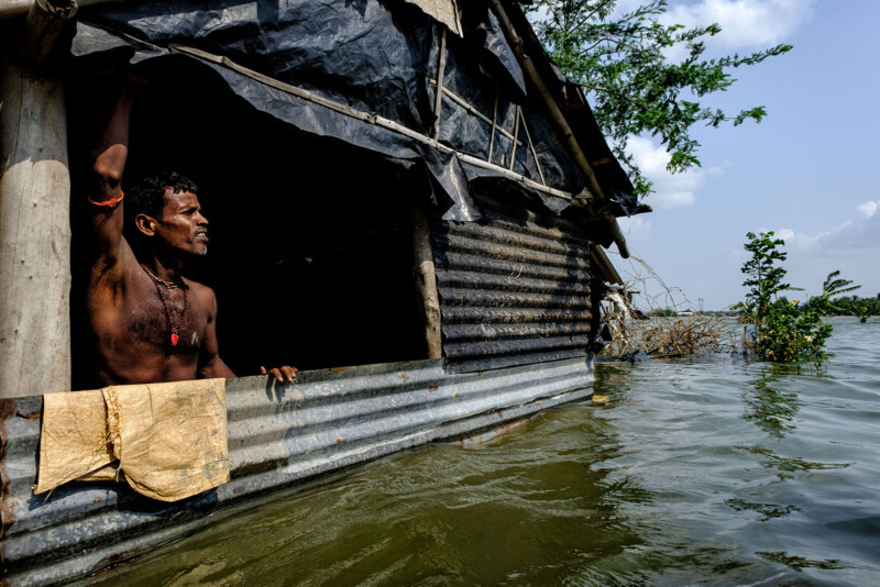 A man stands in a makeshift shelter made of corrugated metal, partially submerged in floodwater. He looks outward, holding onto the structure's frame. The surrounding area is flooded under a blue sky with sparse clouds. Trees partially submerged in water are visible.