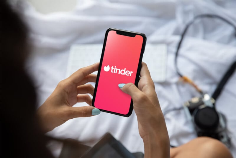 A person holds a smartphone displaying the Tinder app logo on the screen. The background features a white keyboard and a camera placed on a soft, white surface. The person has light blue nail polish.