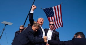 A man in a suit raises his fist in the air while surrounded by security personnel. An American flag is prominently displayed in the background against a clear blue sky. The scene appears animated and energetic, likely capturing a moment of celebration or emphasis.