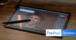 A digital pen rests on a tablet screen displaying a photo editing software with an image of a smiling person wearing a hat. The tablet is placed on a wooden surface. The bottom right corner features the "PetaPixel Reviews" logo.