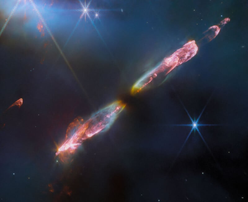 This image of Herbig Haro 211 taken by the James Webb Space Telescope shows a bright, elongated nebula with pink and purple hues against a dark, starry background. Bright stars with diffraction spikes are visible.