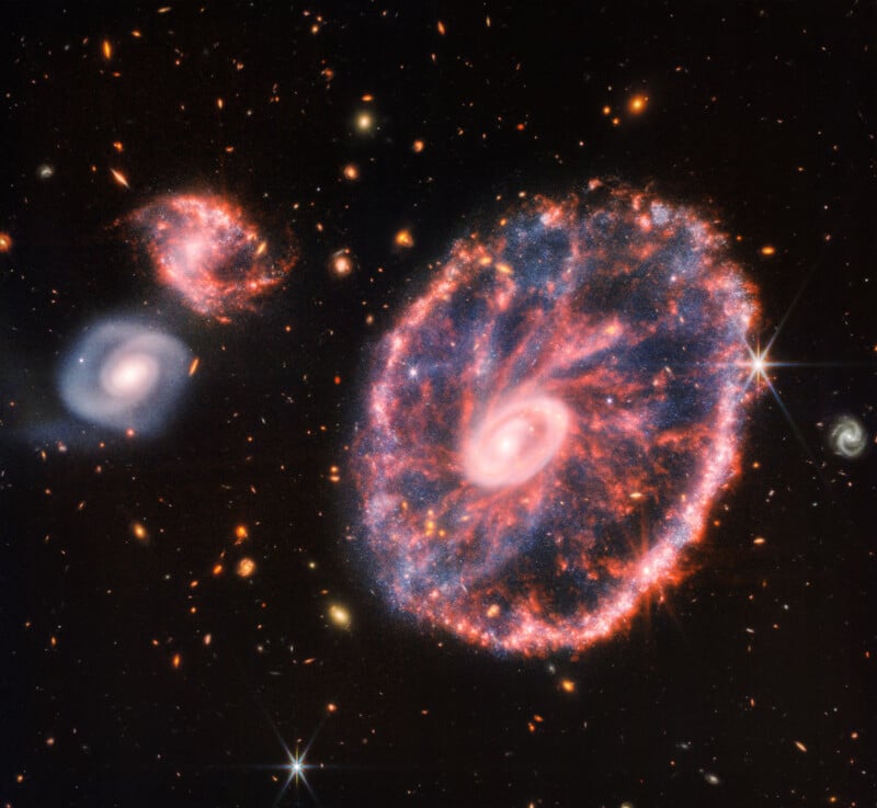 An image of the Cartwheel Galaxy captured by the James Webb Space Telescope shows a bright ring structure with a vibrant, glowing core. Spiral galaxies and other celestial objects surround it, set against the darkness of space sprinkled with stars.