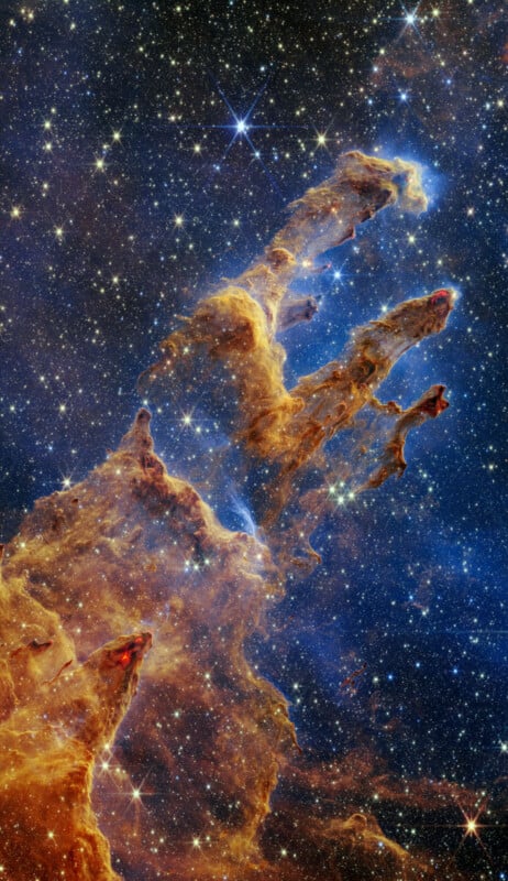 Immagine vibrante dei Pilastri della Creazione nella Nebulosa Aquila, ripresa dal telescopio spaziale James Webb.  Colonne imponenti e semitrasparenti di gas e polvere circondano uno sfondo stellato, mostrando un mix di tonalità blu scuro, rosso e oro.